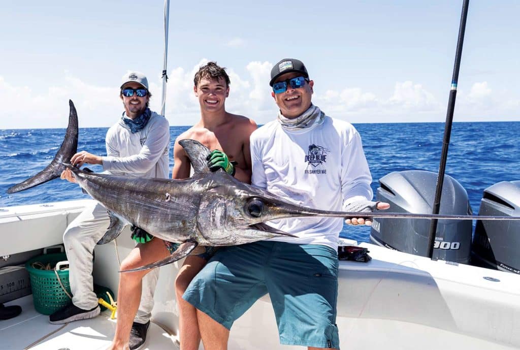 Three anglers sit on a boat deck and hold a large swordfish.