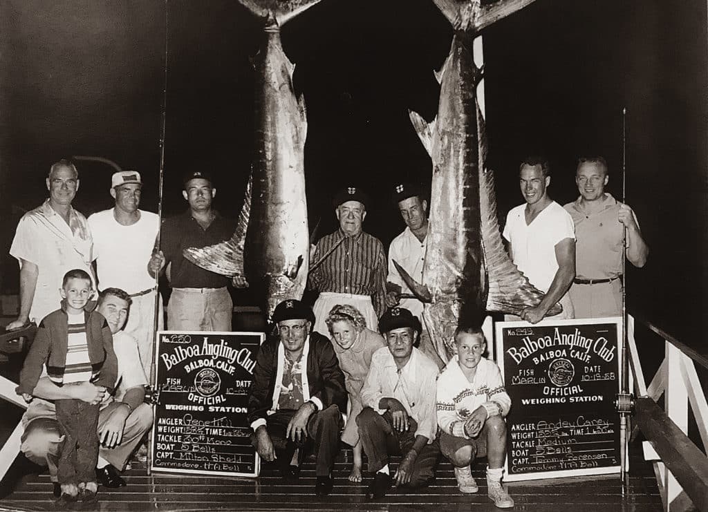 A black and white photograph of a fishing team posing next to two large marlin hanging from hooks on a marine boat deck.