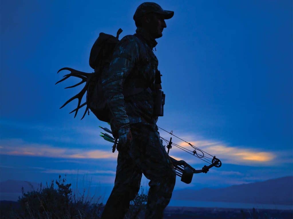 A silhouette of a bowhunter against a dark blue sky at night.