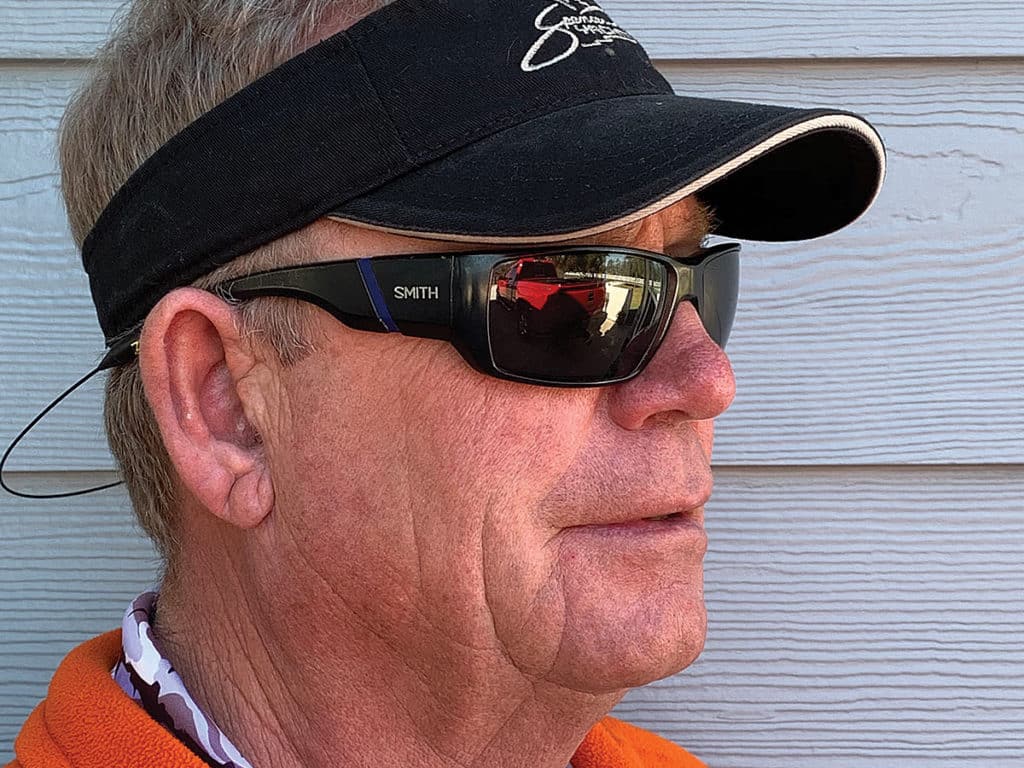 An older man in sunglasses, visor, and orange shirt.
