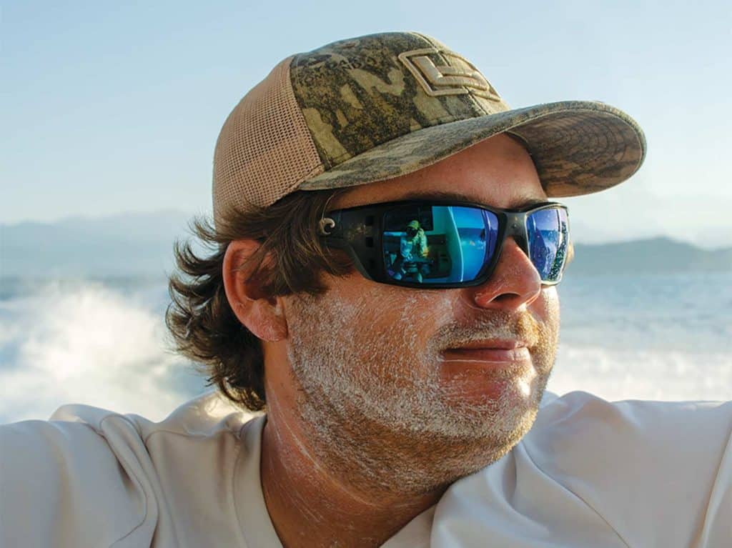 A boat captain sits on a boat deck wearing sunglasses and sunscreen.