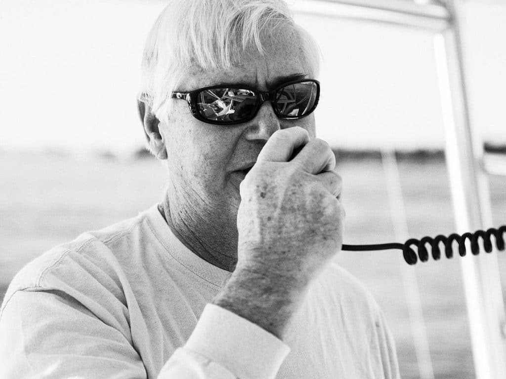 A black and white image of a boat captain at the helm, talking on a two-way radio.