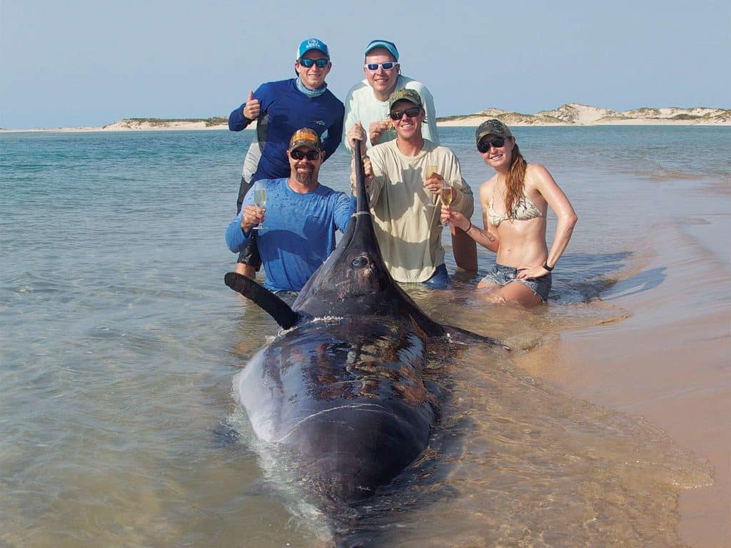 A group of people stand beside a marlin that's been pulled into the shallow waters.
