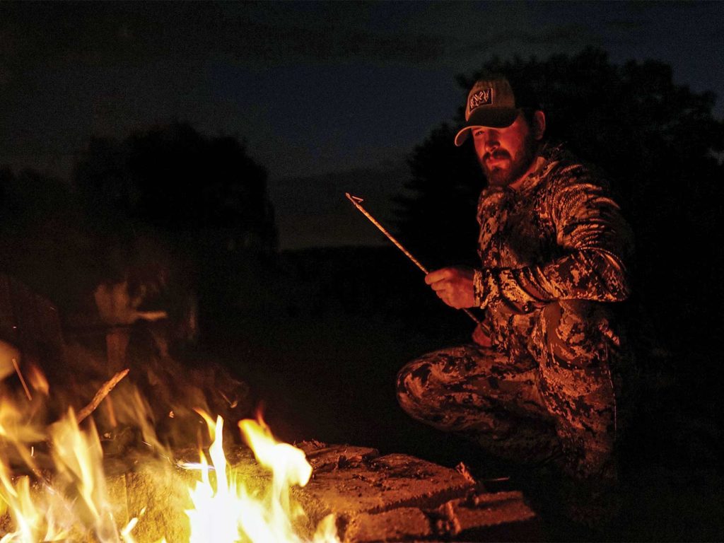A hunter sits by a campfire at night.