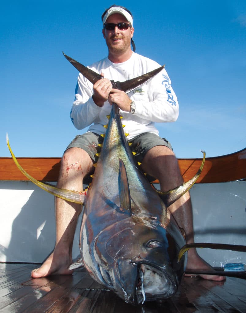 A man sits on a boat deck holding the fin of a large bluefin tuna.