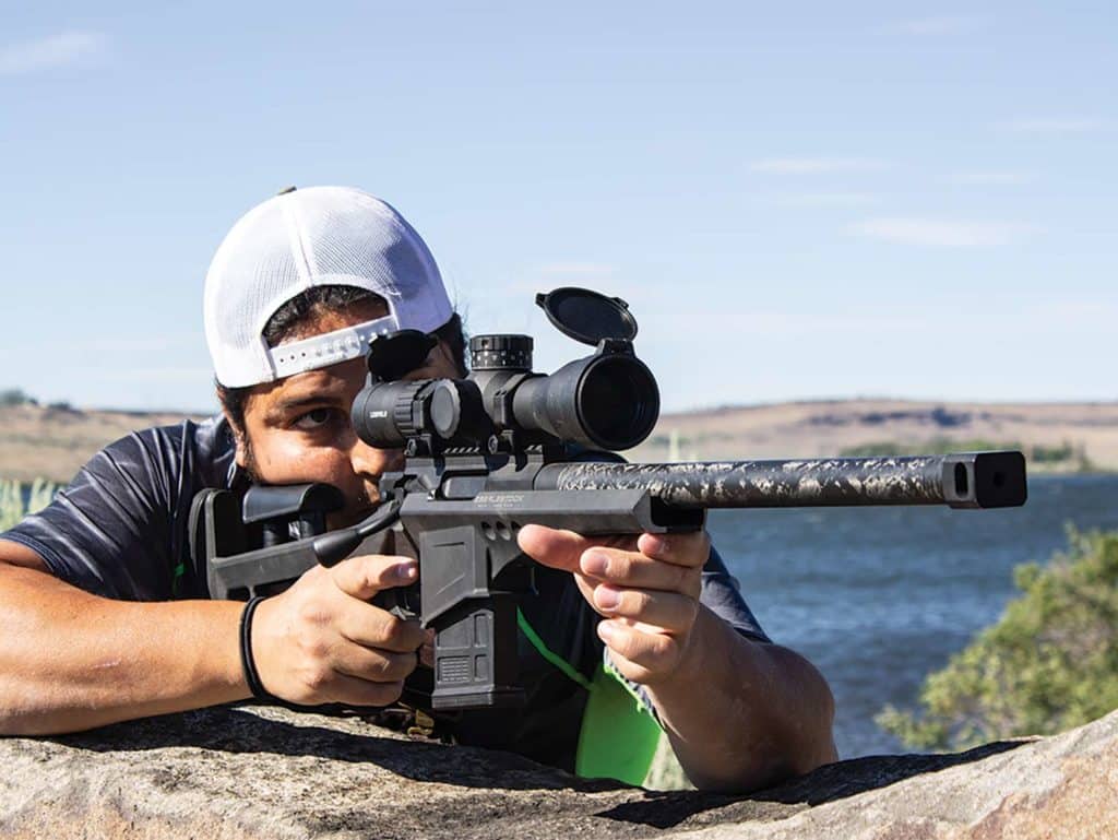 A man aims a scoped rifle while next to a lake.