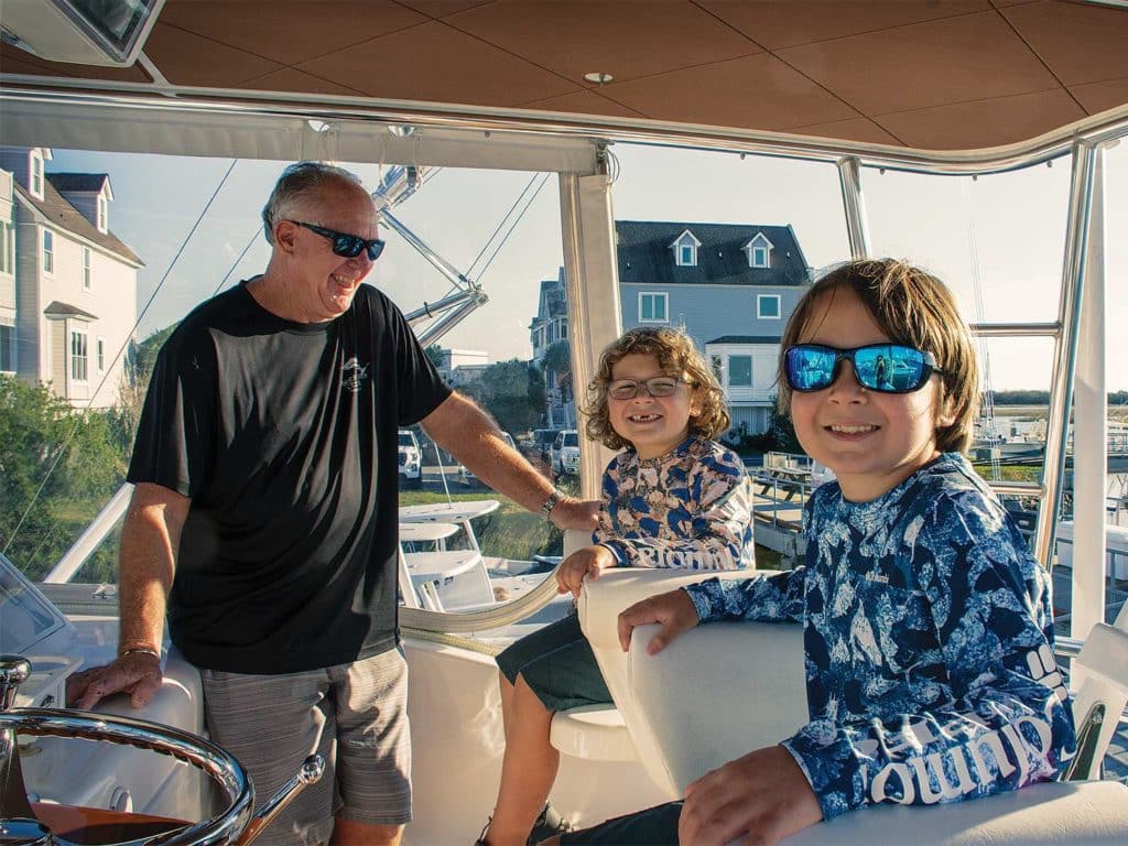 A man stands in a boat helm next to two children.