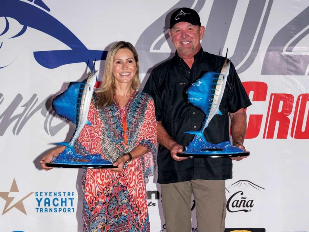 A man and woman stand on an awards stage holding sailfish trophies.