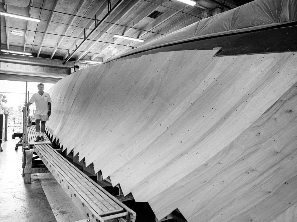 A black and white image of a man standing near a boat hull in a boat building shop.