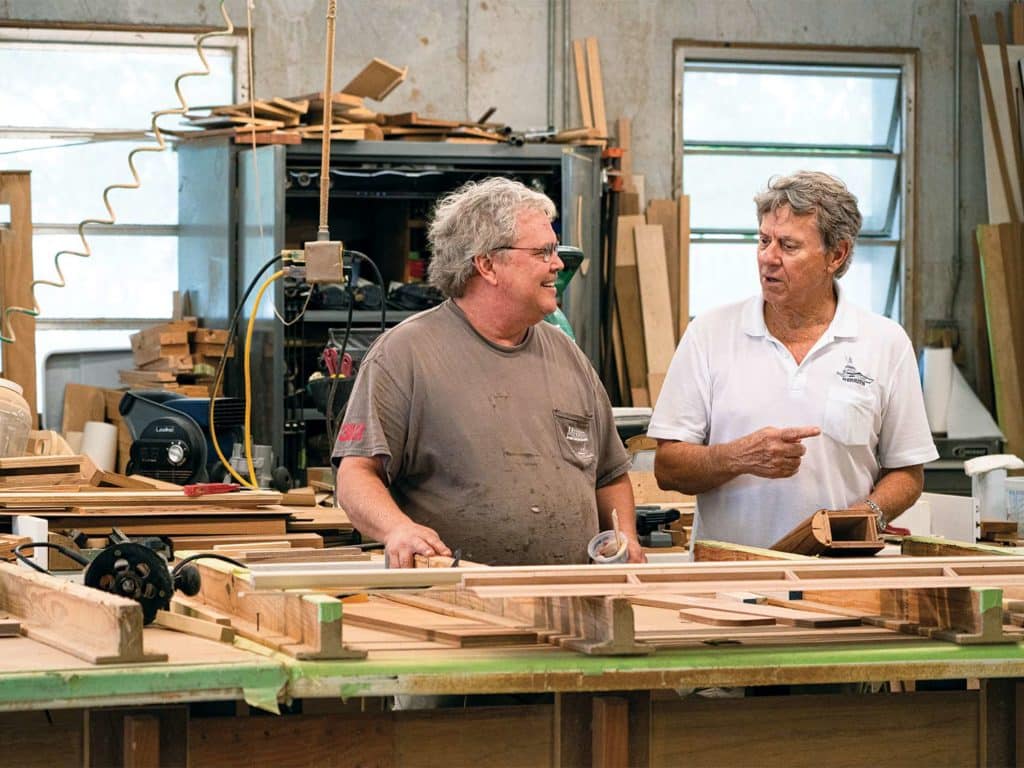 Two men smile and chat at the woodworking desk of a boat building workshiop.