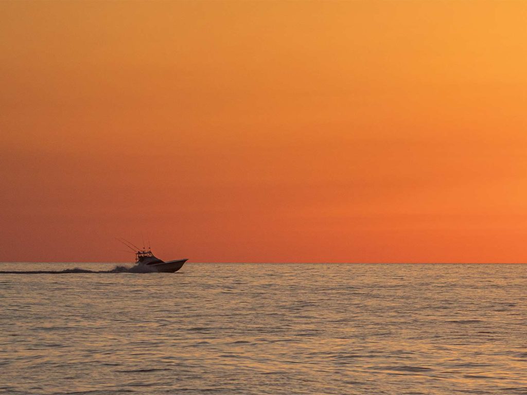 A view of the sunset over the ocean. A silhouette of a sport fishing boat makes its way across the waters in the distance.