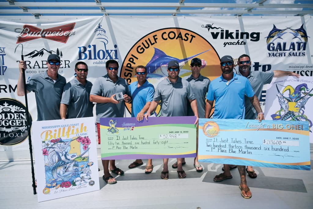 A team of sport fishing anglers pose with giant checks in front of a sponsored event banner.