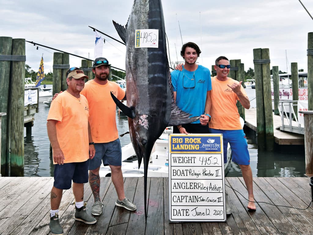Four anglers pose next to a large blue marlin that hangs on a fishing dock at a marina.