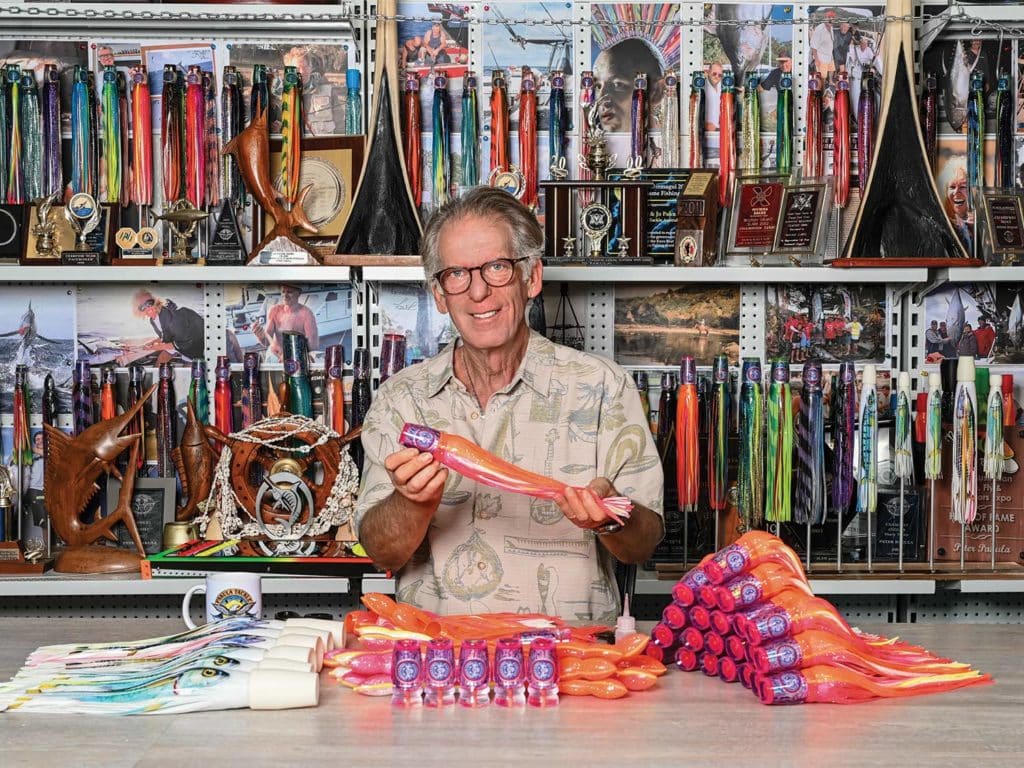 A man stands behind a table, holding a large marlin bait. The wall behind him is full of trophies and awards.