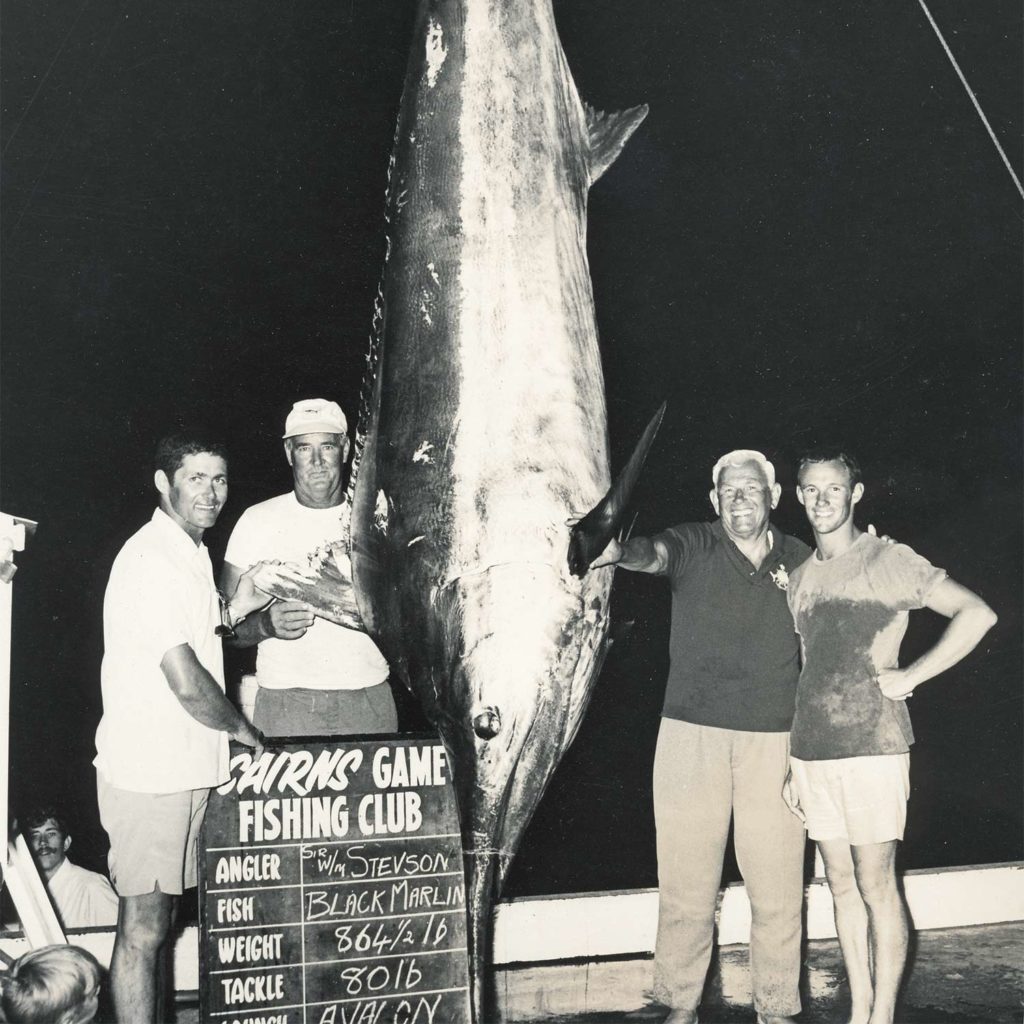 A black and white photo of sport-fishing team standing beside a large blue marlin hanging up for scoring.