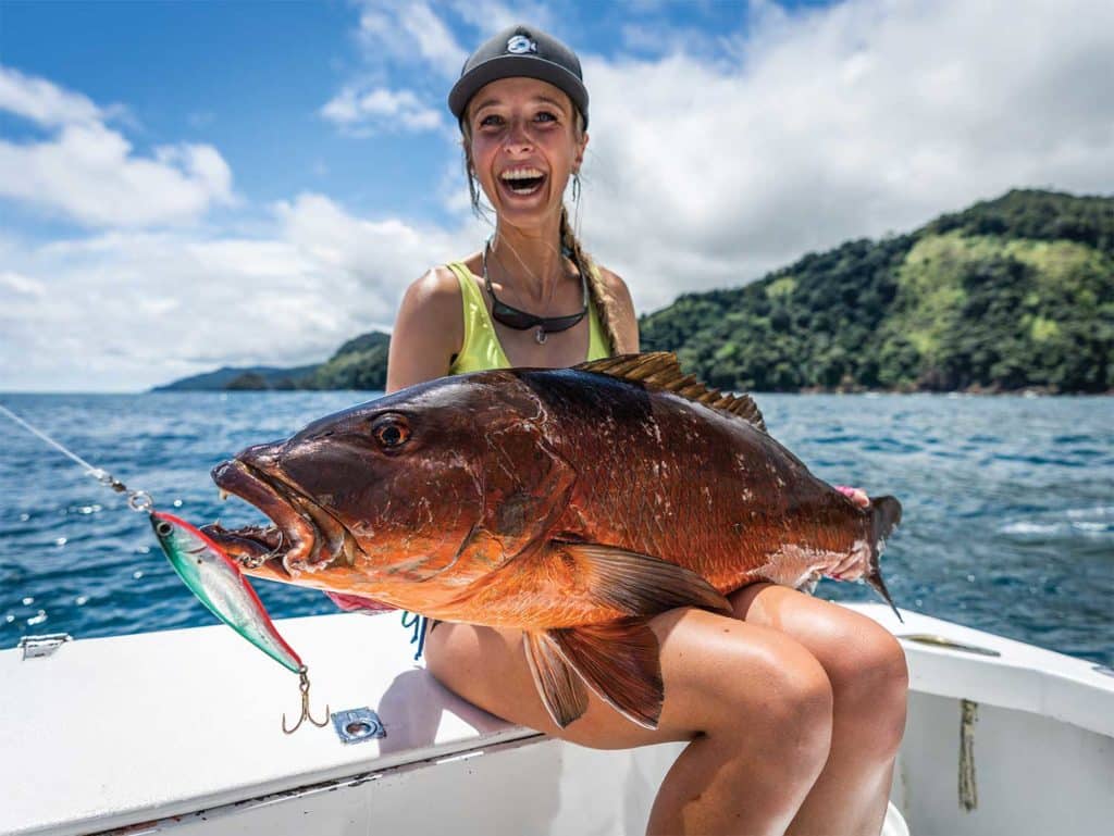 Matilda Leijon, wearing fishing gear and a cap holds up a red cubera snapper. Ocean and a forest island of Panama.