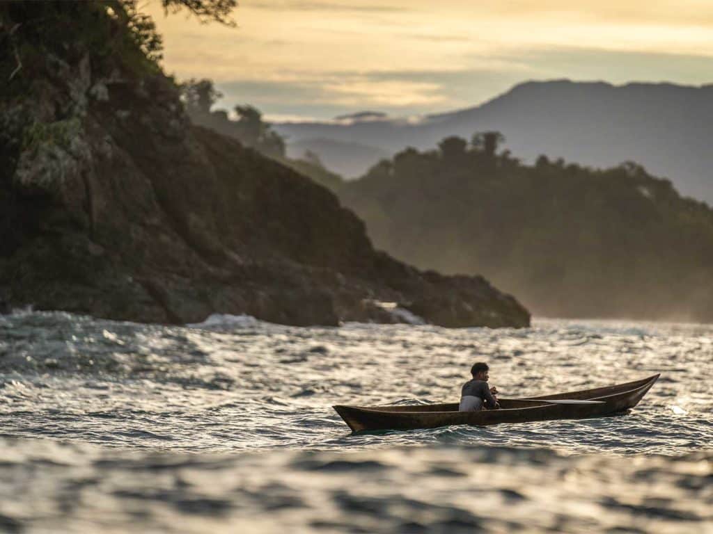 Sun sets over the Panama forest landscape. A single small dinghy with a person inside it floats on the waves.