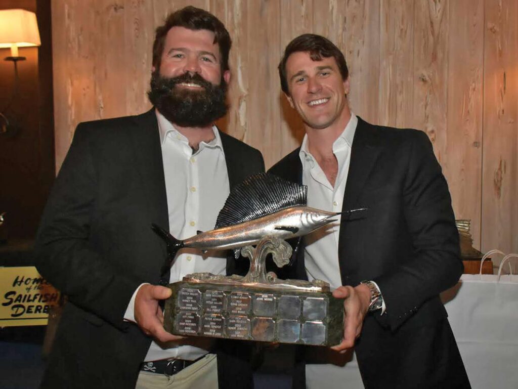 Two men standing and smiling while holding a sailfish trophy between them.
