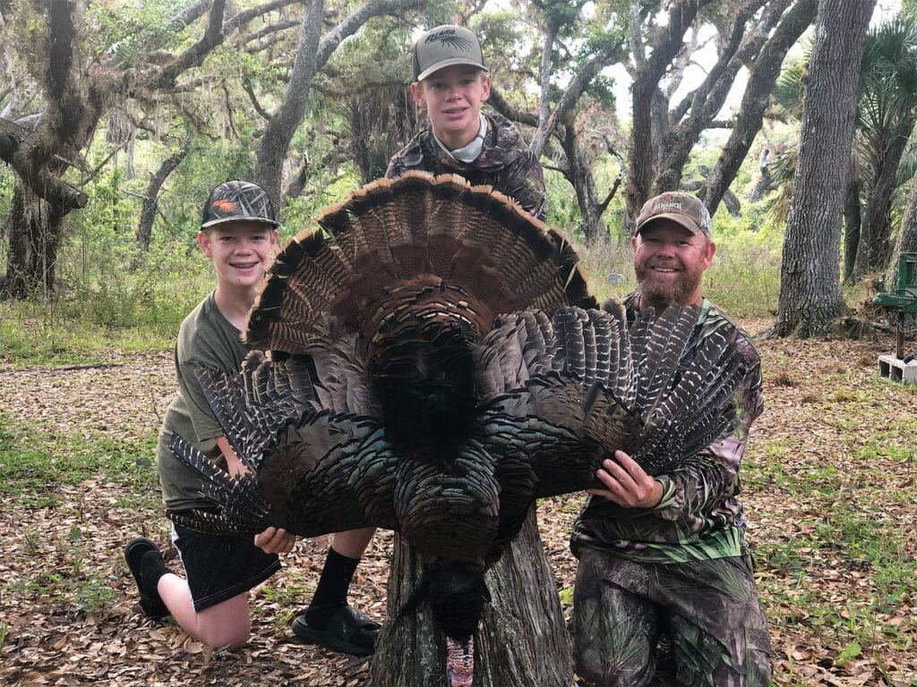 A father and two young sons sitting and posing next to a large turkey in the woods.