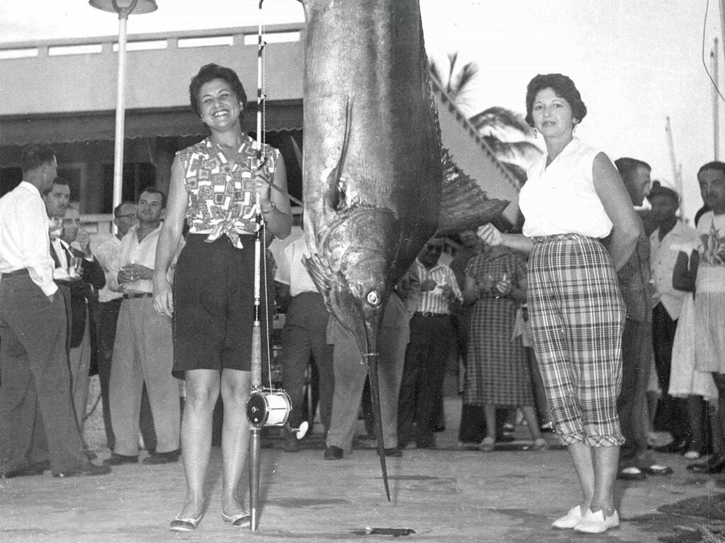 A black and white image of two women standing next to a large blue marlin.