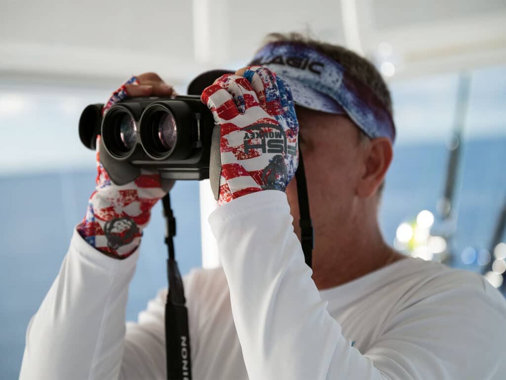 A boat captain views through binoculars while at the helm.