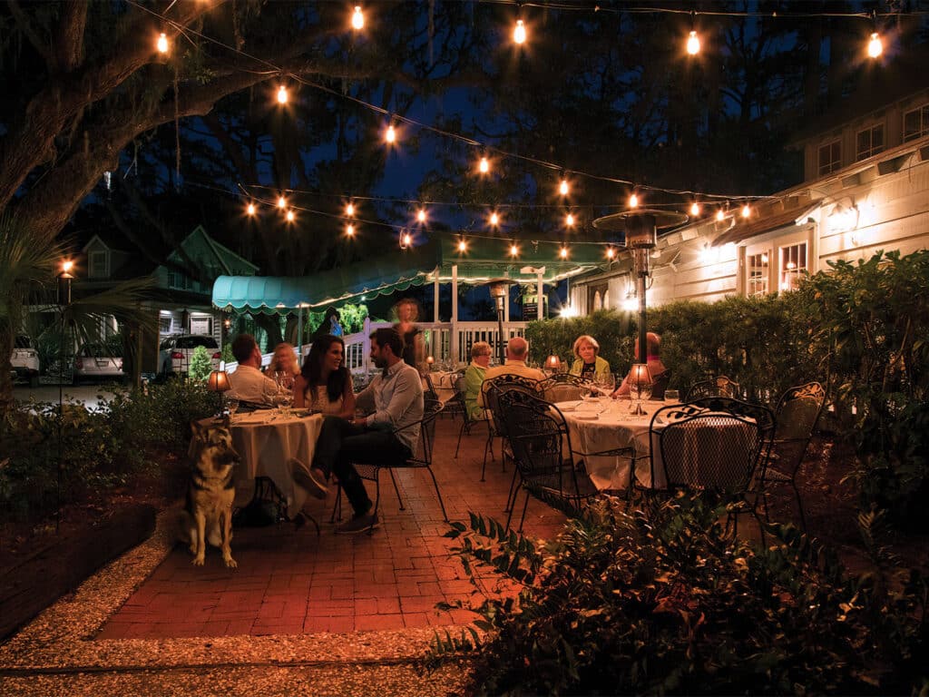 A nighttime image of an outdoors seating area at a restaurant. The patio is full of diners and is lit by overhead string lights.
