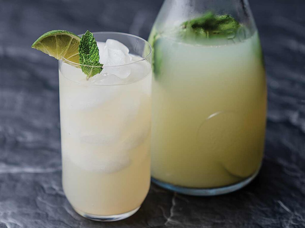 A glass of minty spiked limeade next to a pitcher full of the same cocktail.
