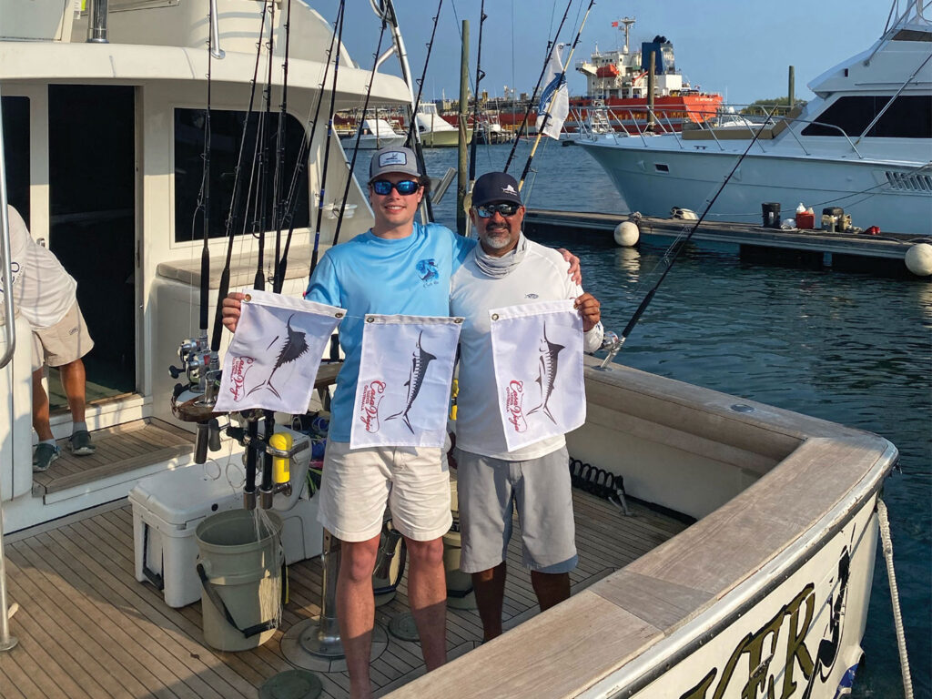 Two men standing in the mezzanine of a sport-fishing boat holding up marlin flags.