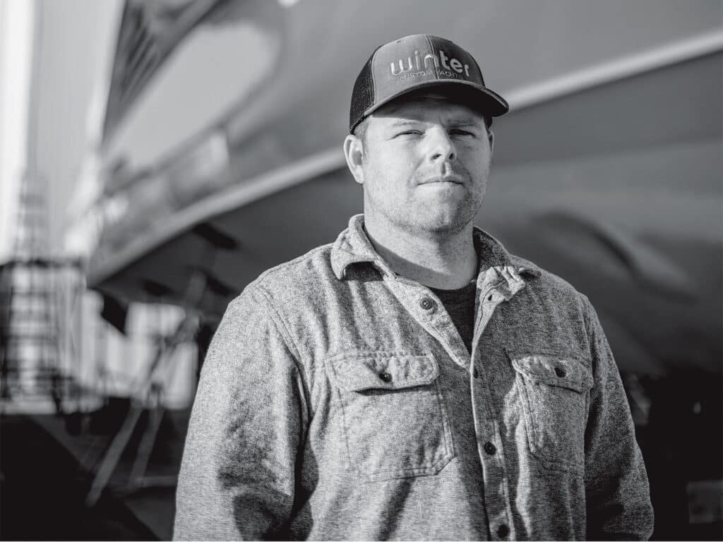 A black and white portrait of Tim Winters standing in front of a custom boat hull.