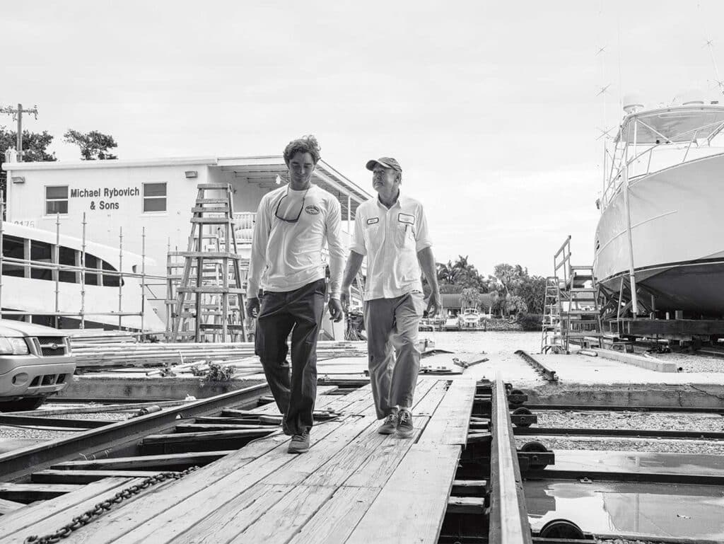 A black and white image of two men walking across the docks of a boat building marina.