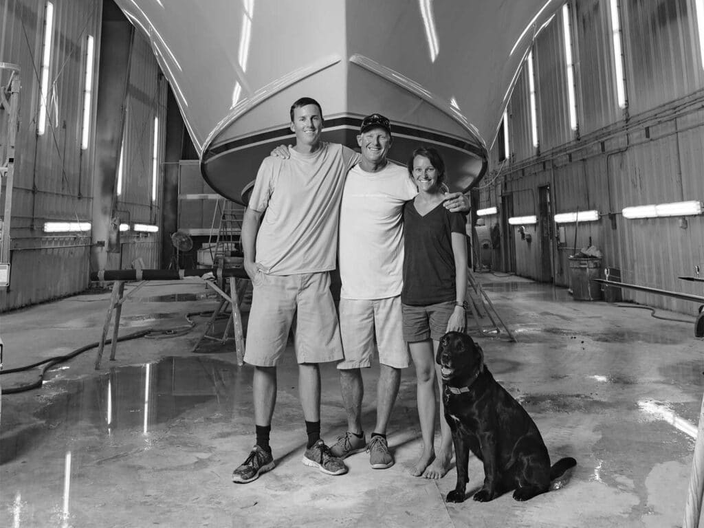 A black and white image of the Bayliss family standing in front of a boat hull with their family dog.