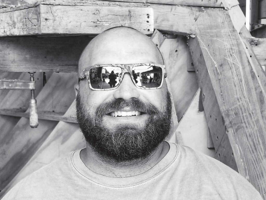 A black and white image of a shaved head, bearded man wearing sunglasses.