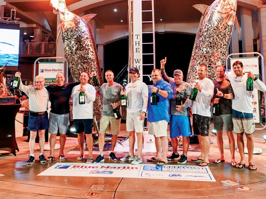 A sport-fishing team of 10 pose and celebrate in front of two marlin metalwork statues that are spouting flame.