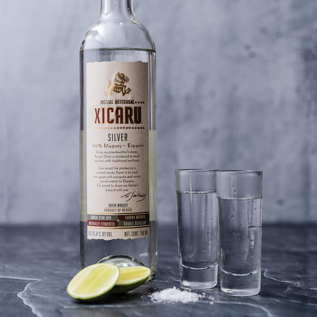 A bottle of Xicaru Silver Mezcal next to two shot glasses and lime wedges.