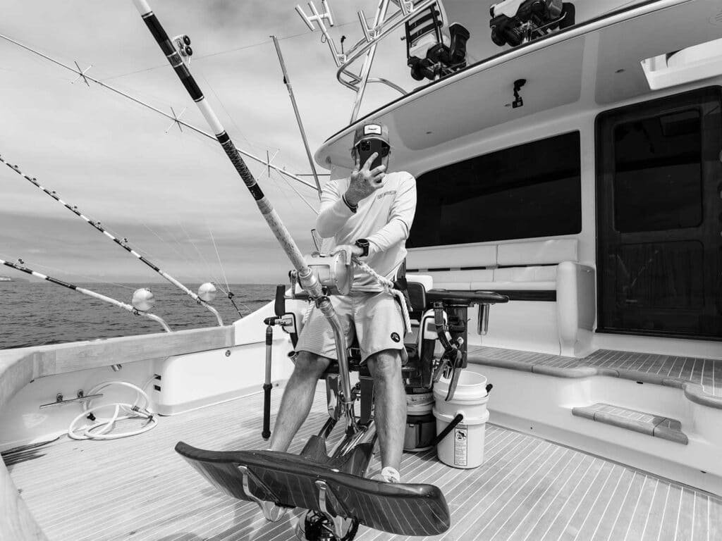 Black and white image of a sport-fishing angler seated in a fighting chair, filming a catch.