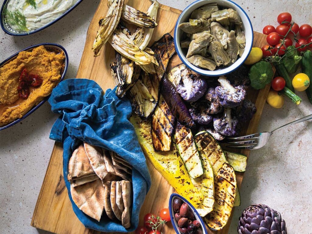 Charred Vegetables with White Bean and Tahini-Yogurt Dips arranged assorted style on a wooden cutting board surrounded by whole vegetables.