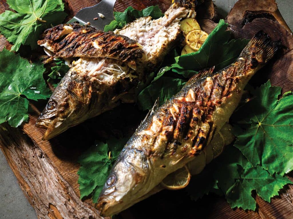 Grill-Roasted Whole Branzino on a wooden block over beds of green leaves.