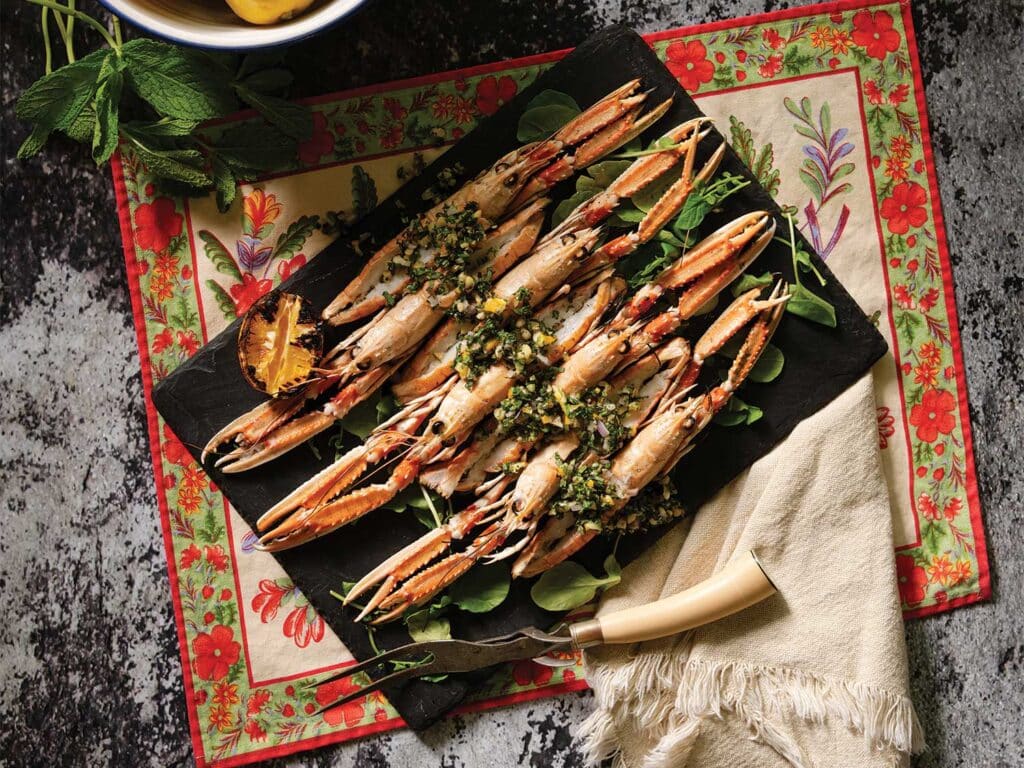 Grilled Butterflied Langoustines with Brown Butter and Pine Nut Gremolata arranged on a serving board over a bed of greens and garnished.