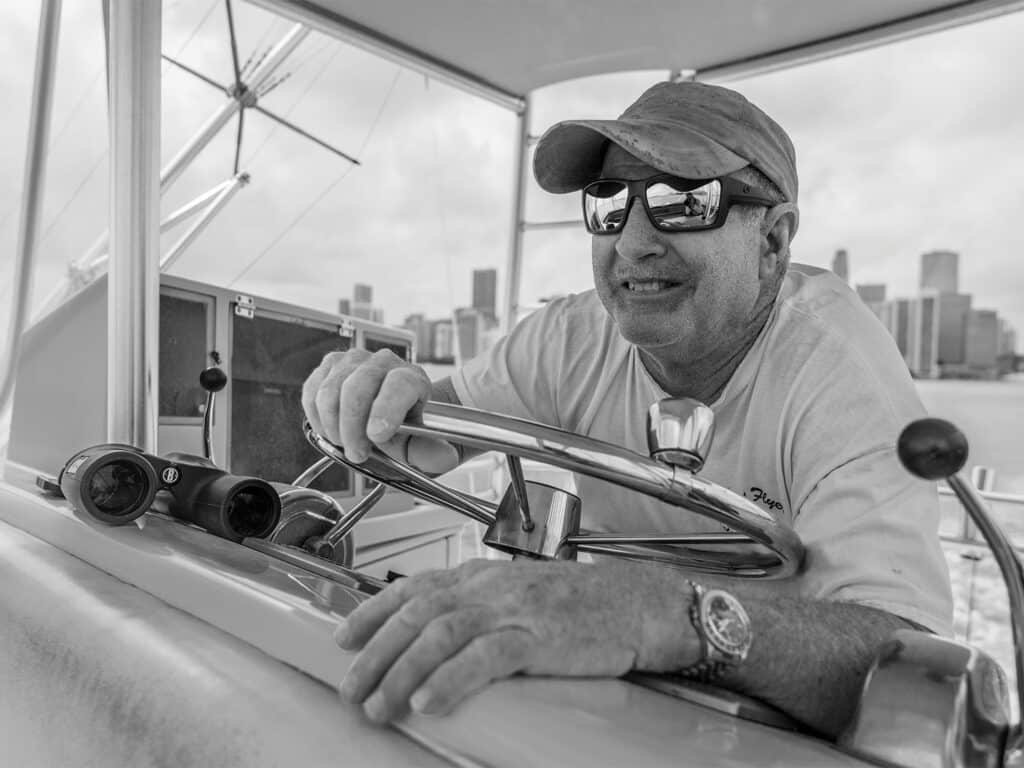 A black and white image of a man wearing a cap and sunglasses at the helm of a sport-fishing boat.