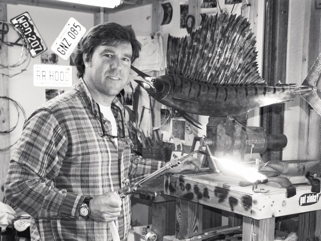 A black and white image of Hanes Hoffman standing in his workshop in front of a sailfish trophy.