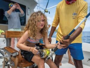A young girl with curly golden hair sits in the cockpit of a sport-fishing boat while the crew assist her in reeling.