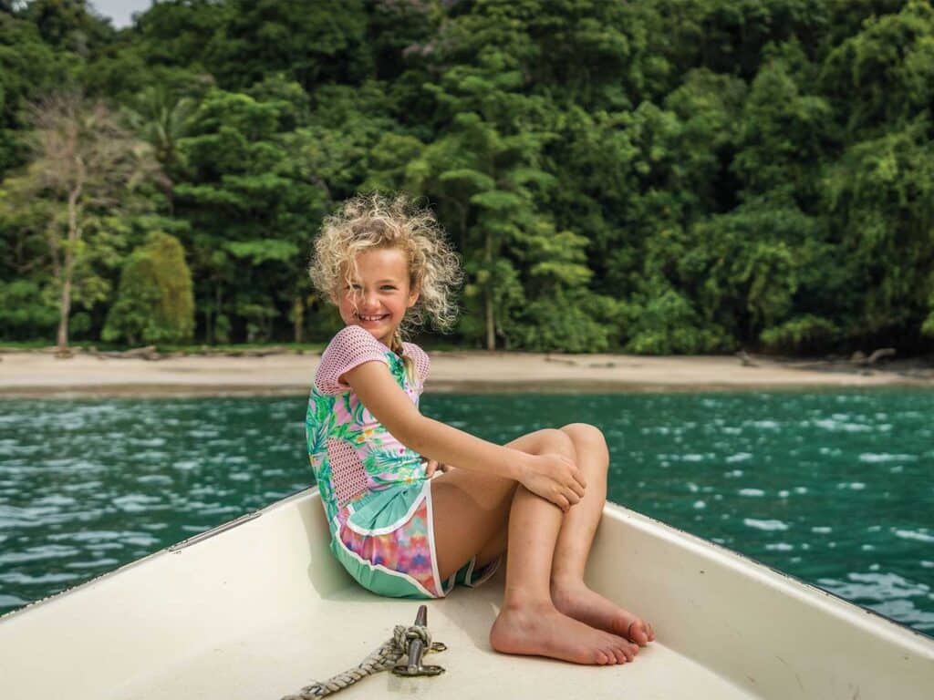 A young girl with curly golden hair sits at the bow of a boat in front of a tropical backdrop.