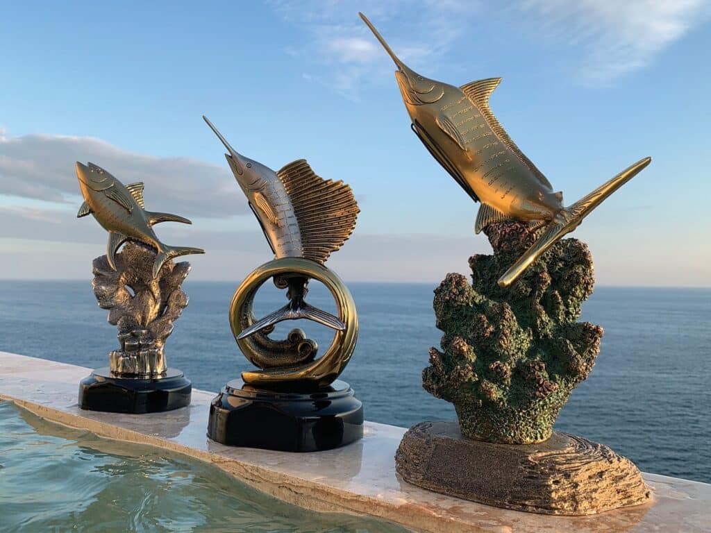 Three sport-fishing tournament trophies arranged on a wooden beam with the ocean in the background.