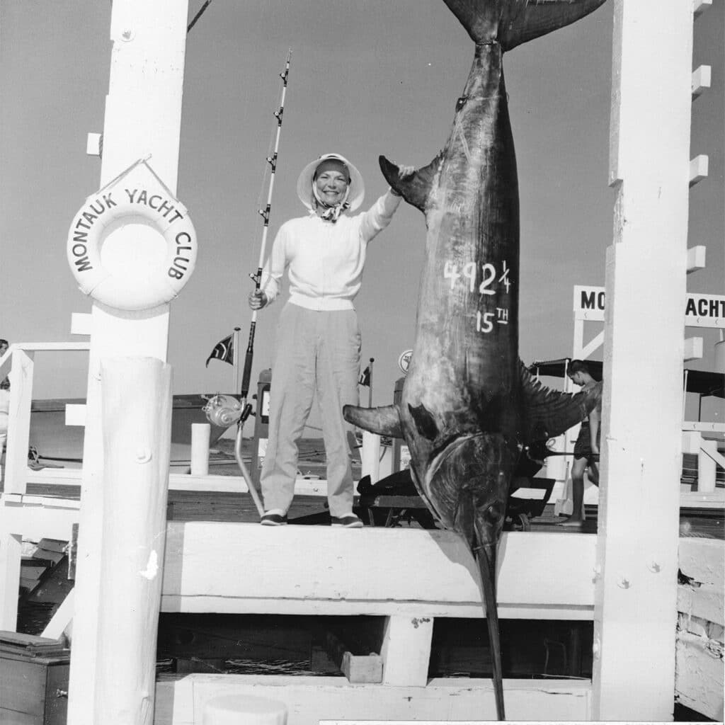 A black and white image of Dorothea Cassullo standing next to a large swrodfish suspended by dock rigging.