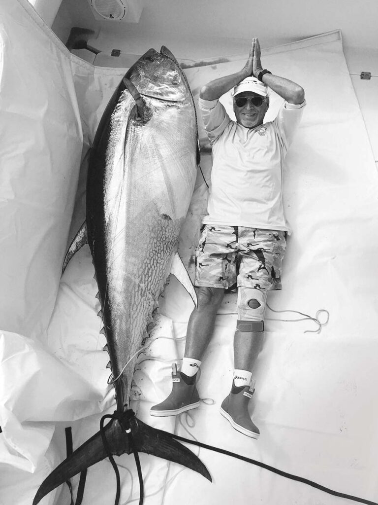 Black and white image of Jimmy Buffett laying next to a large bluefin tuna to show off its massive size, which length is more than Buffett's height.