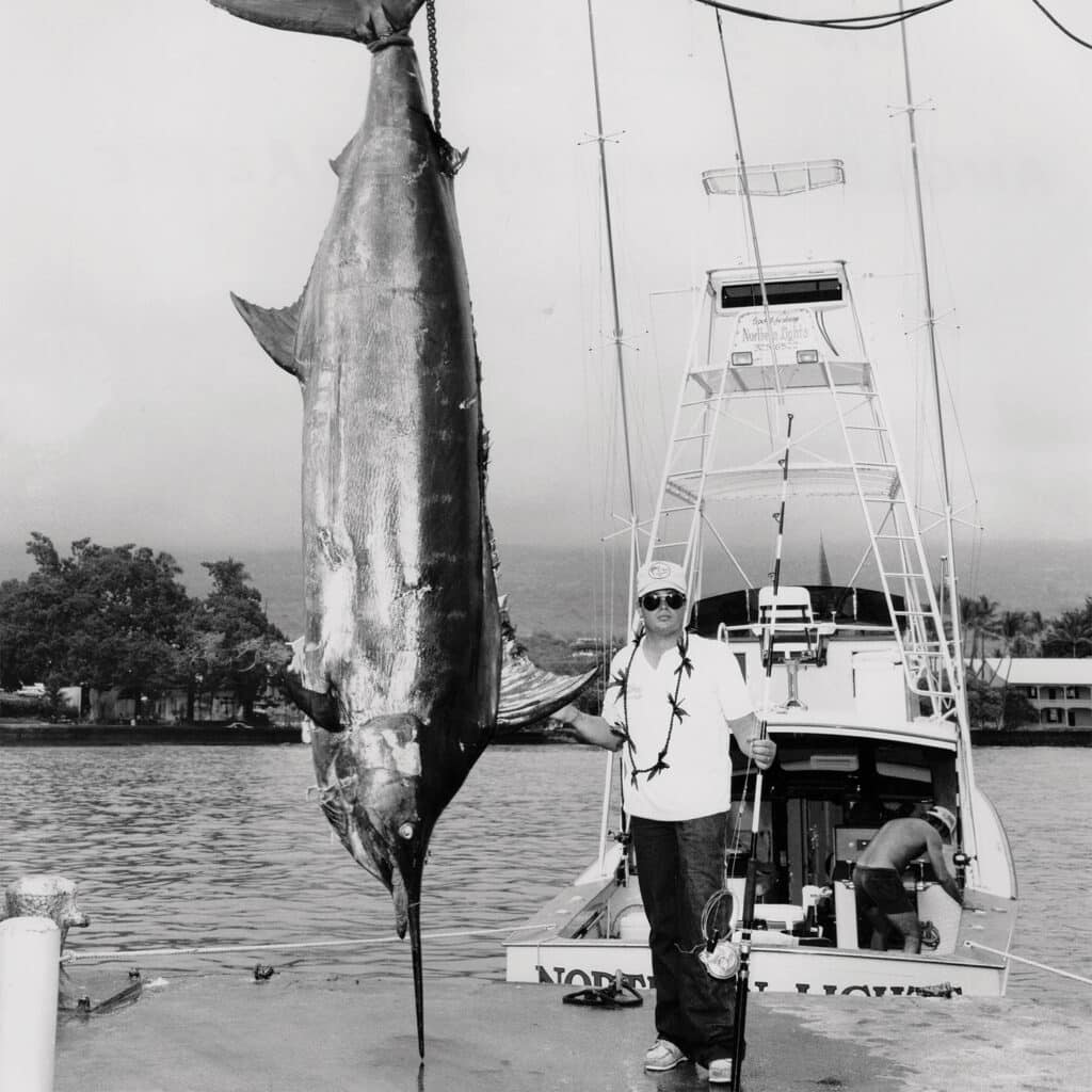 A black and white image of Kelley Everette standing next to a grander Pacific blue marlin suspended from dock rigging.