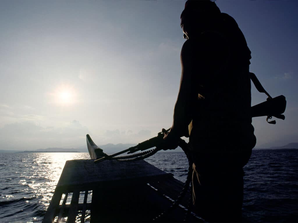 Silhouette of a man holding a gun while standing on a boat, overlooking the ocean horizon.