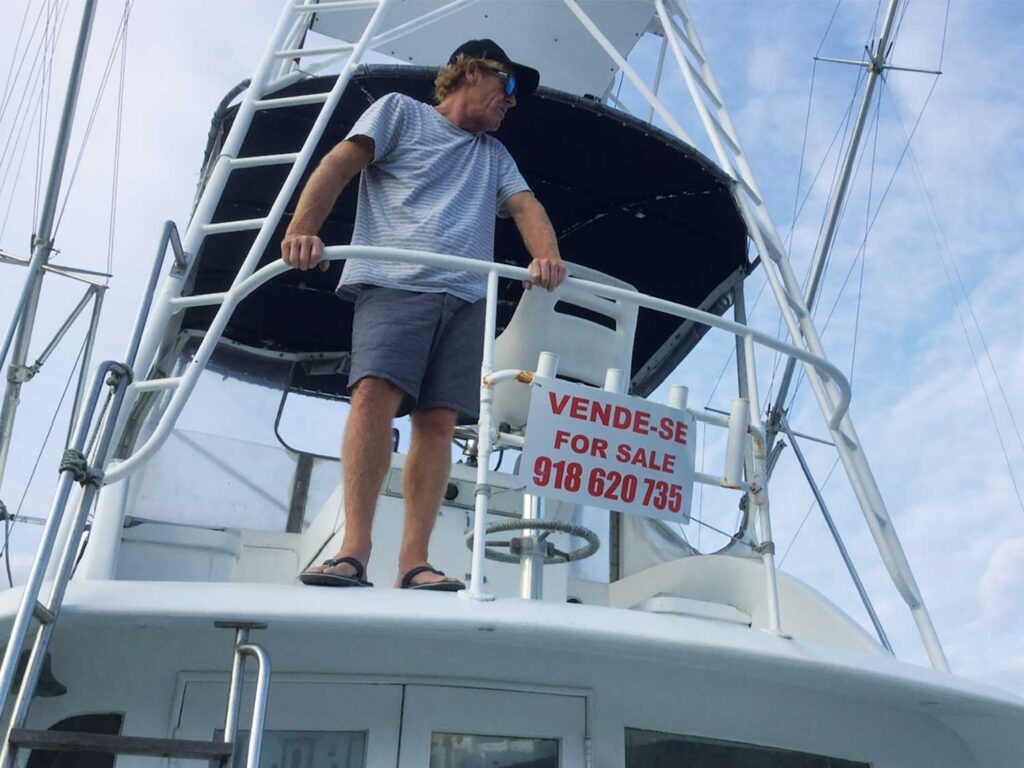 A man stands in the tower of a sport-fishing boat with a sale sign on its railings.