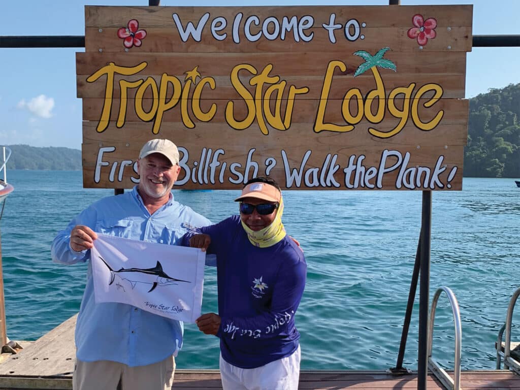 Two men hold a marlin flag while standing in front of the Tropic Star Lodge sign.