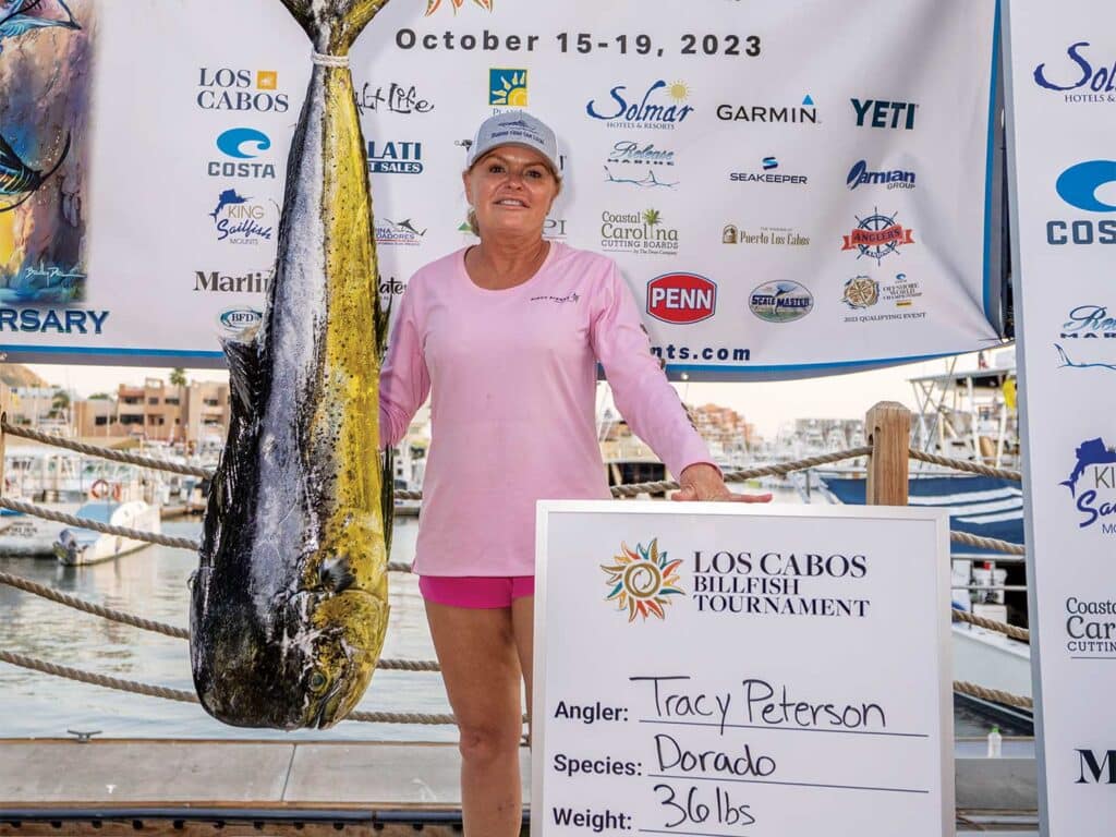A woman in a pink shirt stands next to a prized dorado gamefish.
